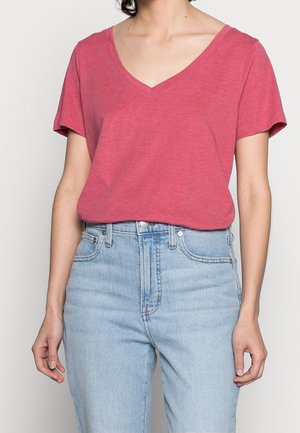 Person iført en rød V-hals t-shirt med korte ærmer, som er stoppet ned i lyseblå, højtaljede jeans, med hænderne afslappet ved siden.