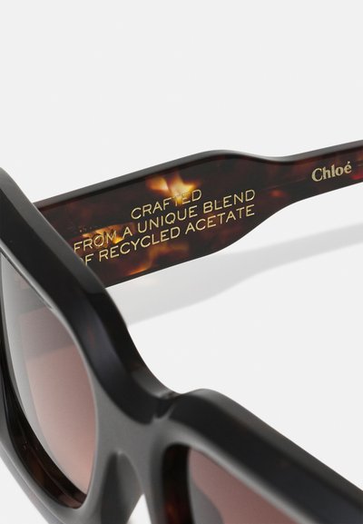 Okulary przeciwsłoneczne z czarną oprawą i zausznikami w wzór skorupy żółwia, z złotym napisem o recyklingowanym materiale acetatowym i nazwą marki Chloé.