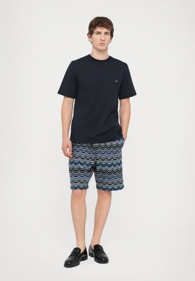 Marinblå t-shirt, mönstrade shorts med blå och gröna zickzack-mönster, och svarta loafers. Shortsen har resårmidja.