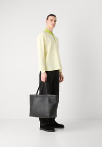 Bolso tote de cuero negro con doble asas, textura suave, forma rectangular. Modelo vistiendo una blusa amarilla clara con cuello verde y jeans oscuros.