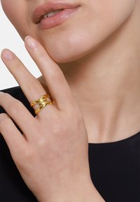 Gouden ring met een gedraaide, in elkaar grijpende ontwerp. Soepele, glanzende textuur. Draagt op de wijsvinger tegen een donkere achtergrond.