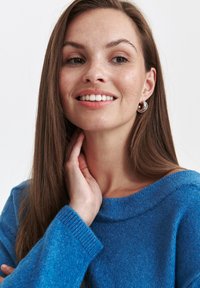 Niebieski sweter z dzianiny o szerokim dekolcie i prążkowanych mankietach. Modelka nosi srebrne kolczyki w kształcie kół i ma długie, proste włosy.