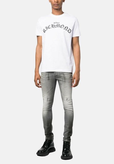 T-shirt bianca in cotone con grafica "John Richmond" in nero, abbinata a jeans grigi slim-fit e stivaletti neri con lacci.