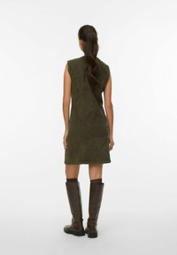 Khaki groene mouwloze jurk met een getailleerde vorm, voorzien van een split aan de achterkant en een onafgewerkte zoom. Gepaard met bruin leren laarzen tot de knie.