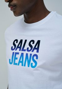 Camiseta de algodón blanca con el texto "SALSA JEANS" en un degradado de azul oscuro a azul claro. Cuello redondo, textura suave, ajustada al cuerpo.