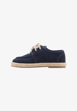 Zapato de ante azul marino con cordones crema y suela de esparto beige. Presenta una puntera redondeada y un diseño de perfil bajo.