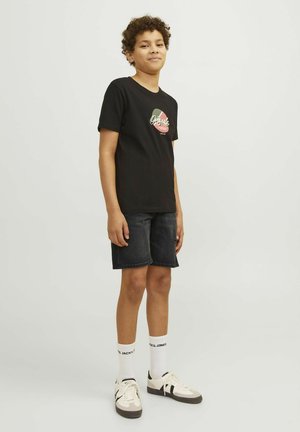 Tricou negru cu imprimeu grafic, purtat cu pantaloni scurți din denim închis. Modelul poartă șosete albe cu logo și adidași bej cu accente negre.