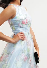 Vaaleansininen kukkamekko, jossa on vartalonmyötäinen yläosa ja levenevä helma. Mekossa on sileä pinta sekä kirkkaan pinkkejä ja vihreitä kukkakuvioita.