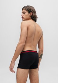 Schwarze Boxershorts mit einem roten "HUGO"-Bund, die auf einem Modell von hinten mit glatter Textur und enganliegendem Design präsentiert werden.