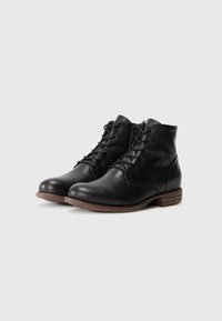 Botines de cuero negro con un diseño de punta de cap, que presentan cordones y una cremallera lateral, acabados con una suela marrón texturada.
