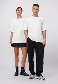 Camisetas blancas de gran tamaño con la palabra "STUDIOS" estampada. Modelo femenino lleva una falda mini de cuero negro y botas de tobillo; modelo masculino en pantalones negros y zapatillas.