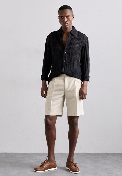 Cămașă neagră, texturată, cu butoni, pantaloni scurți bej plisati și pantofi de bărbați maro. Design simplu, cu culori și materiale contrastante.