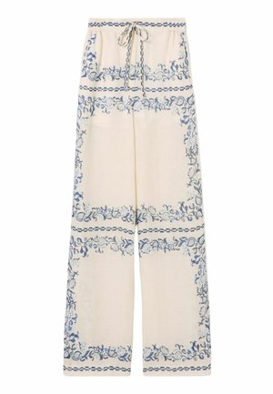 Femme aux cheveux attachés en chignon, portant un gilet crème boutonné, un pantalon large crème avec des motifs floraux bleus, des sandales marron et un sac à main rayé sur l'épaule.