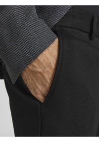 Svarta byxor med en figursydd design, med en sidficka. Manschetten på en mörkt mönstrad skjorta är synlig, vilket visar en strukturerad tyg.