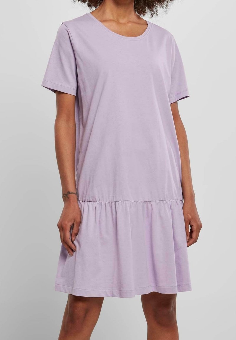 Personne portant une robe lavande ample à manches courtes avec un détail de volants froncés au-dessus du genou, debout sur un fond uni.