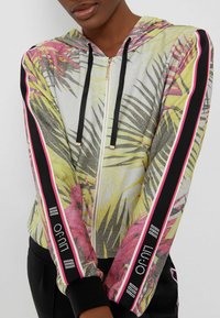 Capuchon zipjack met een metallic glans, voorzien van kleurrijke tropische bladeren in geel en roze, zwarte manchetten en decoratieve strepen.
