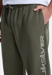 Olivgröna mjukisbyxor i mjukt tyg med elastiskt midjeband och dragsko. Har sidofickor och en vit logotyp tryckt längs benet.