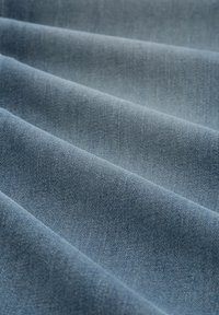 Tkanina dżinsowa w jasnoniebieskim kolorze, charakteryzująca się gładką fakturą z diagonalnym splotem skośnym i delikatnymi różnicami w kolorze na fałdach.