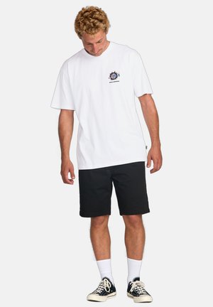 White cotton t-shirt with a graphic logo on the chest, paired with black shorts. Model wearing black sneakers and white socks.