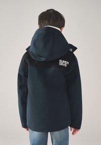 Tumesinine jope kapuutsiga, mustad õlaaktsendid ja valge logo tagaosas. Materjal näib olevat sile ja struktuurne disain.