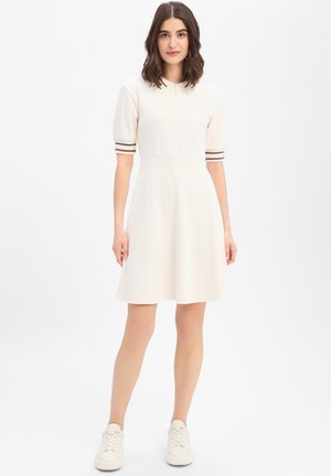 Jonge vrouw met donker schouderlang haar, die een crème korte mouwen jurk draagt met gestreepte boorden en witte sneakers.