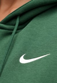 Zelená mikina s kapucňou z mäkkej látky s textúrovaným povrchom. Prednú časť zdobí biele logo Nike a kapucňa obsahuje šnúrky na stiahnutie.