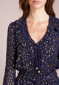 Marinblå klänning med guldigt blommönster, volangkrage och knytning fram. Har långa ärmar och en smockad midja.