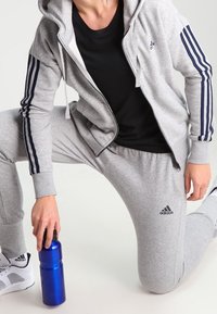 Tuta grigio con strisce blu, felpa con zip e pantaloni jogger. T-shirt nera sotto. Bottiglia d'acqua blu in mano, sneakers bianche visibili.