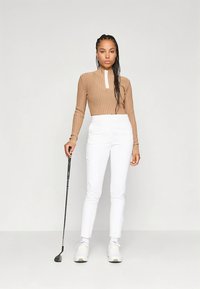 Lichtbruine geribbelde top met lange mouwen en een witte ritssluiting, gecombineerd met witte, op maat gemaakte broek en witte sneakers, en een golfclub vasthoudend.