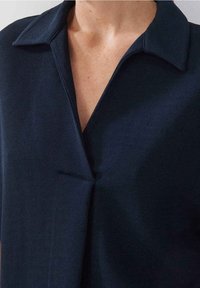 Chemise bleu marine en tissu slub, avec col en V, détails de coutures verticales et un léger décolleté en V, mettant en avant un design propre et moderne.