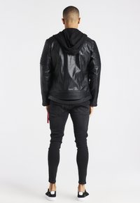 Giubbotto di pelle nera con spalle e schiena a coste, abbinato a jeans neri slim e sneaker nere, con un cappuccio sotto il colletto.