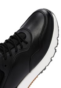 Zwarte leren sneaker met een gestructureerde zwarte mesh-tong, zwarte veters en een witte rubberen zool met een lichtbruine accent.