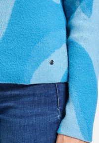 Blauer Strickpullover mit verschiedenen Farbtönen, strukturiertem Finish und schwarzem Logodetail am Saum, kombiniert mit dunkelblauen Jeans.