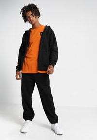 Sudadera con cremallera negra sobre una camiseta naranja, combinada con pantalones de chándal negros y zapatillas blancas; diseño simple; tejido texturizado.
