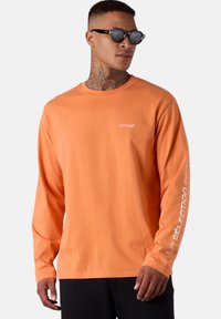 Langärmliges orangenes Baumwoll-T-Shirt mit Rundhalsausschnitt. Mit weißem Logo auf der Brust und Textmuster entlang des Ärmels. Schwarze Hose.