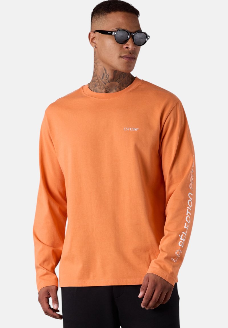 Langärmliges orangenes Baumwoll-T-Shirt mit Rundhalsausschnitt. Mit weißem Logo auf der Brust und Textmuster entlang des Ärmels. Schwarze Hose.