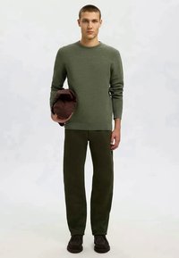 Maglione lavorato a maglia verde con scollatura rotonda, abbinato a pantaloni dritti verde scuro. Il modello tiene in mano una borsa di stoffa marrone. Scarpe marroni completano il look.