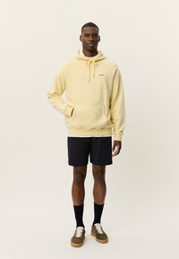 Άνδρας που στέκεται φορώντας ανοιχτόκίτρινο φούτερ με κουκούλα, μαύρο σορτς, μαύρες κάλτσες και καφέ αθλητικά παπούτσια σε απλό ανοιχτό φόντο.