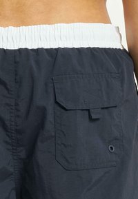 Marineblå shorts laget av lett materiale, med en elastisk hvit midje og en baklomme med klaff og knappelukking.