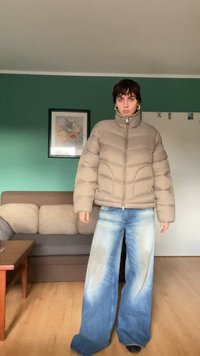 Tan puffy jacket with a high collar and zip, paired with wide-leg, light blue jeans featuring a gradient wash. Indoor setting with green wall.