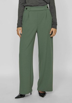 Pantaloni palazzo in tessuto verde scuro con pince, dotati di due bottoni in vita. Abbinati a scarpe con tacco a punta nere.