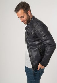 Zwarte leren motorjack met ritssluitingen, textuurbalans op de schouders en een strakke pasvorm, gedragen over een grijs T-shirt en jeans.