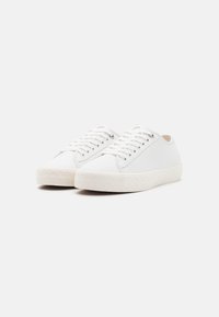 Pari valkoisia, matalia nahkakenkiä, joissa on valkoiset nauhat ja kuvioidut valkoiset kumipohjat, yksinkertaisella vaalealla taustalla.