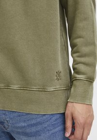 Olive green sweatshirt made of textured fabric, featuring ribbed cuffs and hem, with an embroidered logo detail on the front.