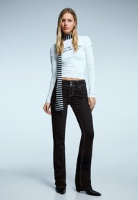 Fekete, bővülő szárú farmer fehér, kontrasztos varrással, hozzáillő, testhez simuló, fehér hosszú ujjú felsővel és fekete-fehér csíkos sállal.