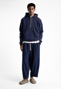 Sweat à capuche bleu marine avec poche avant et cordons de serrage, associé à un pantalon bleu marine ample. Le mannequin porte des chaussures à enfiler beiges.