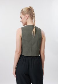 Femme blonde avec queue de cheval tressée portant un gilet sans manches vert olive et un pantalon noir taille haute, vue de dos sur fond uni.