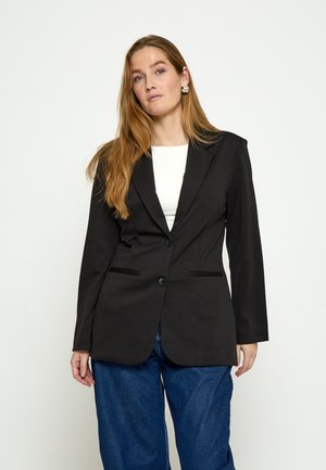 Kobieta z długimi rudymi włosami, ubrana w czarną marynarkę na białej bluzce i niebieskie dżinsy, stojąca na tle jednolitego jasnego tła.