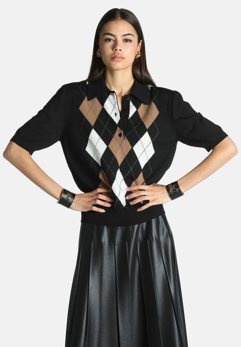 Jeune femme debout, mains sur les hanches, portant un pull argyle noir à manches courtes avec des losanges marron et blancs et une jupe en cuir plissée noire.