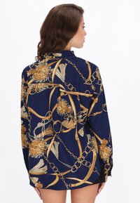 Kvinde med langt brunt hår iført en marineblå bluse med mønstre af guld kæder og kvaster, stående foran en ensartet hvid baggrund.